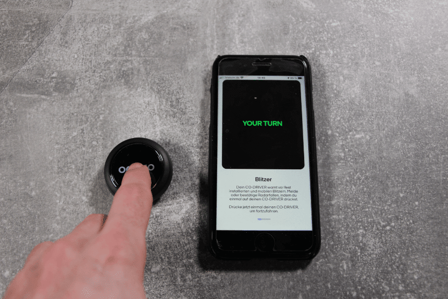 Der Ooono Co-Driver No1 liegt neben dem Smartphone, wird gedrückt und es wird erklärt, wie Du andere vor Blitzern warnen kannst